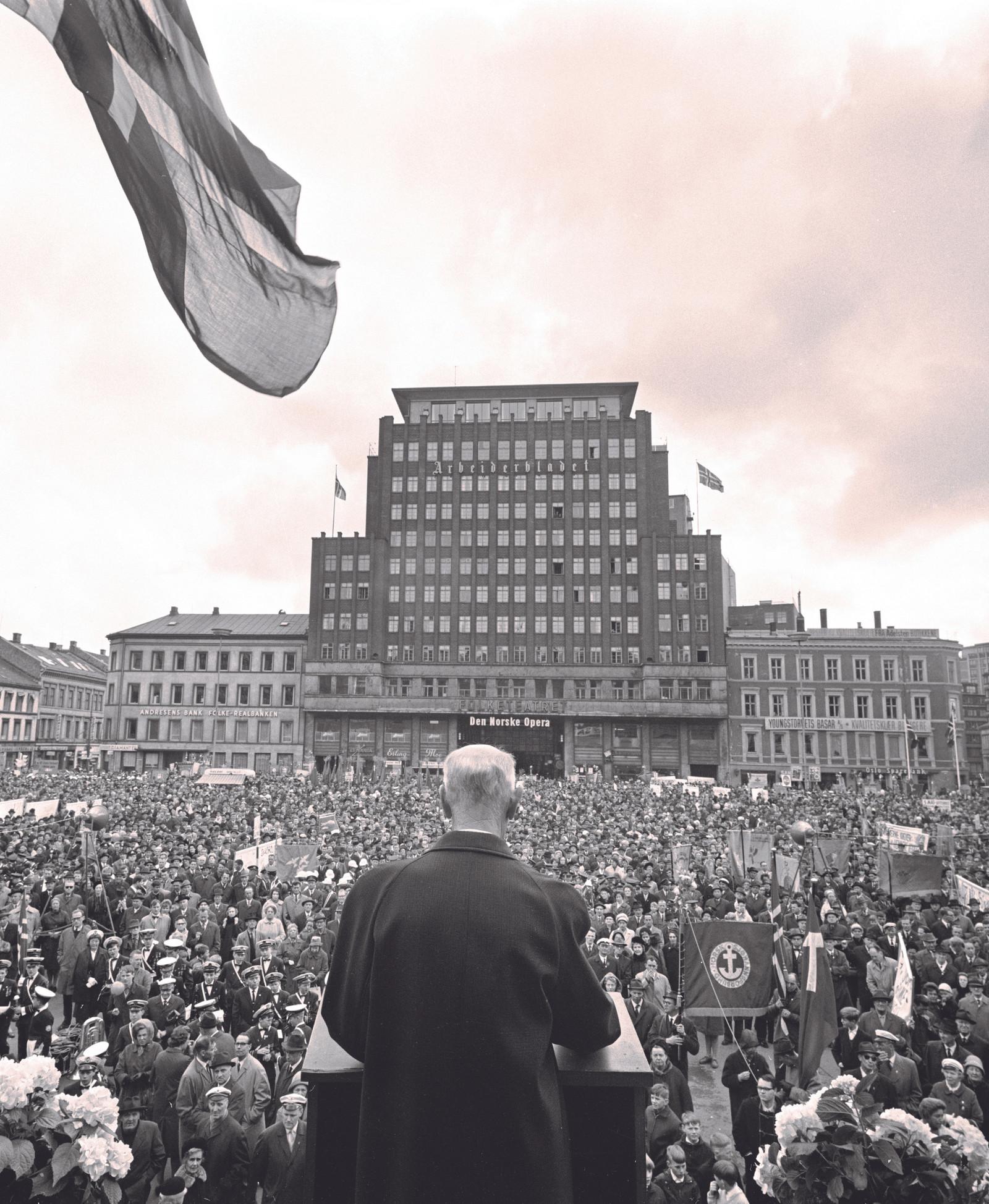 Einar Gerhardsen under vaiende norsk flagg på talerstolen på Youngstorget 1967, sett bakfra.