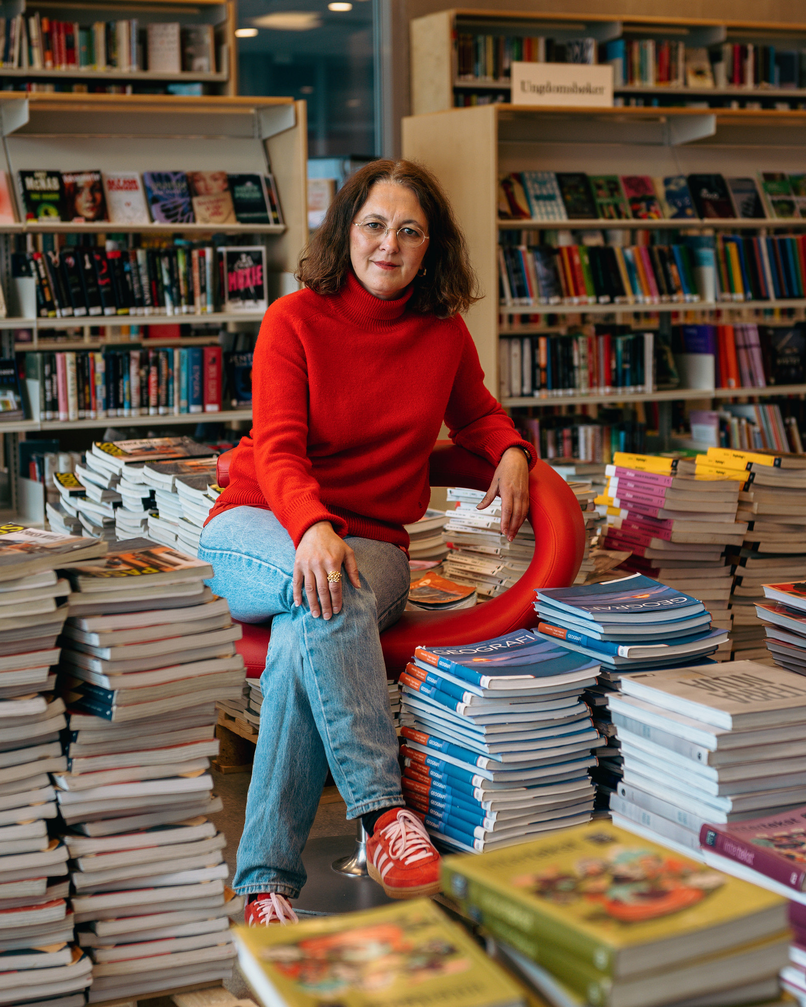 Ane Christiansen med rød genser på rød stol blant en haug med lærebøker